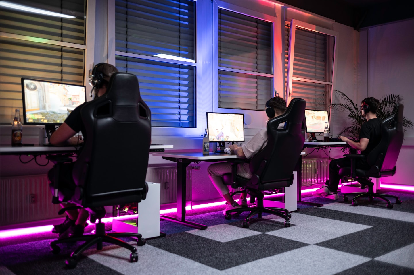 Tres personas jugando videojuegos en computadoras en una habitación luminosa.