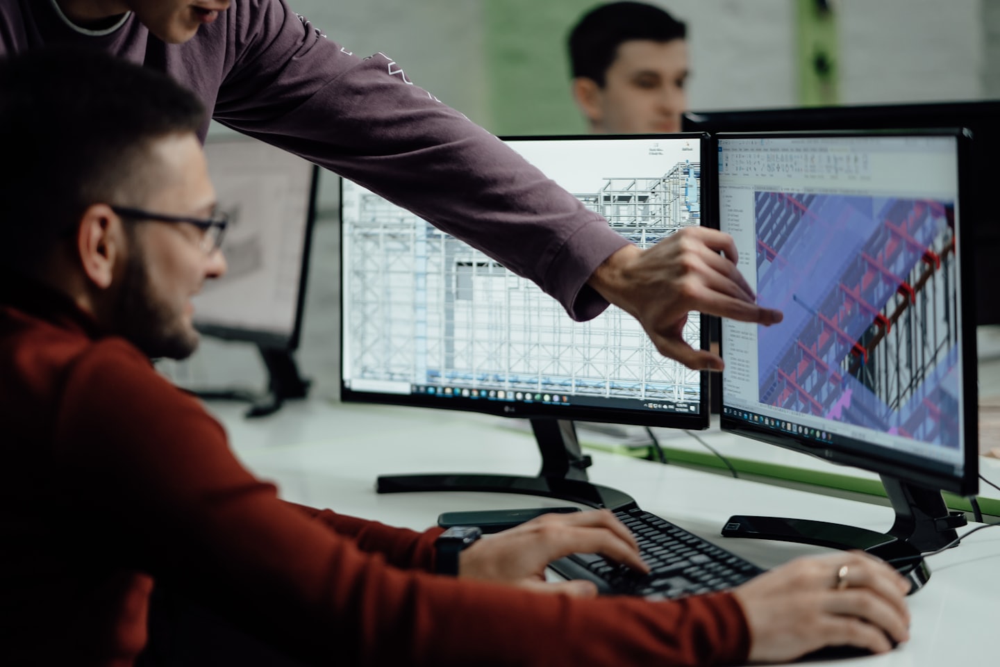 Hombre señala un diseño 3D en monitores duales mientras un colega escribe.
